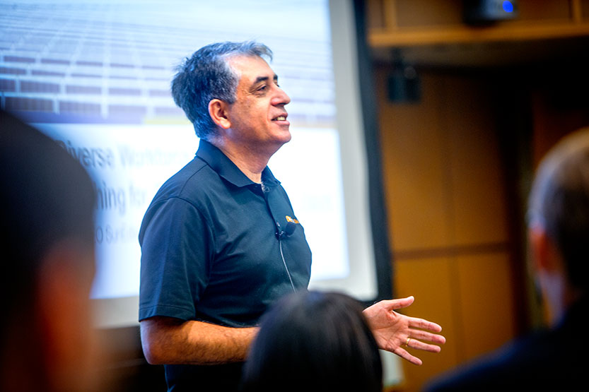 Professor teaching at the front of a classroom with students surrounding him and a screen in the background