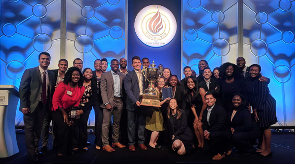 A large group stands on a stage with their trophy