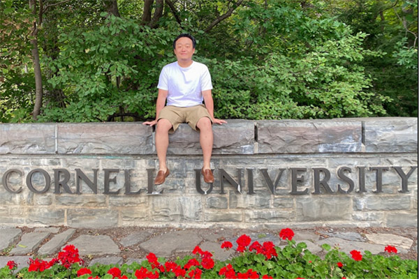 A picture of Tony Tsao sitting on a stone wall with the words Cornell University written on it and trees in the background.