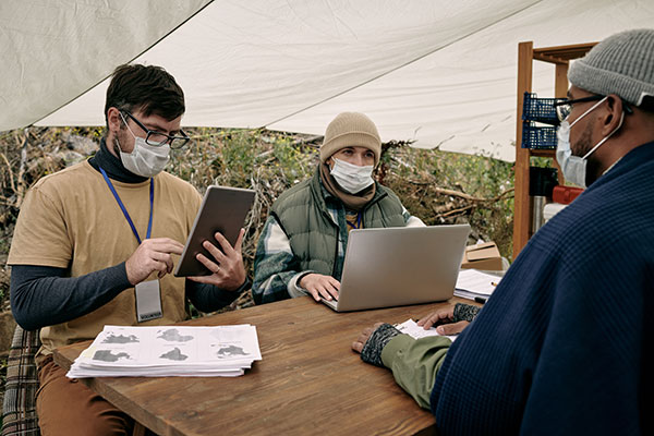 one woman and two men sitting outside at a table conferring and working with a laptop and tablets