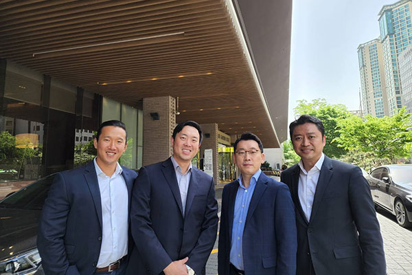four Korean mean in suits standing side by side and smiling with a building and sky and trees in the background