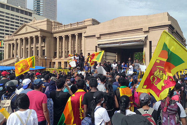 A large crowd of people, some waving Sri Lanka’s flag, facing government buildings and listening to speakers standing in front of those buildings.