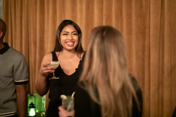 South Asian woman holding a cocktail and smiling with woman blurred in foreground