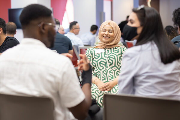 Woman in hijab smiling while listening to people in foreground who are blurred.