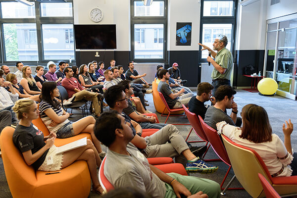 A man holding a microphone speaks in front of students sitting in colorful chairs.