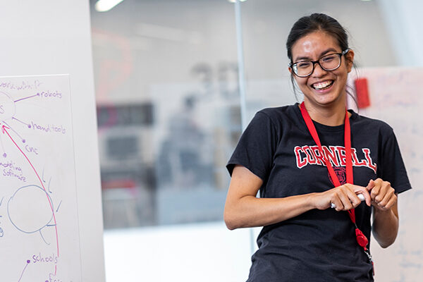 A student wearing a Cornell t-shirt smiles and holds a dry-erase marker in front of a whiteboard.