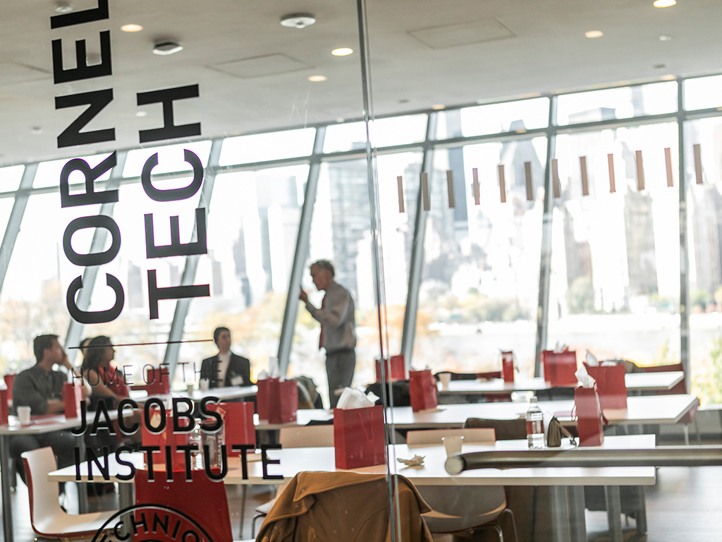 A glass wall labeled with “Cornell Tech” in the foreground and a group of people inside the room in the background.