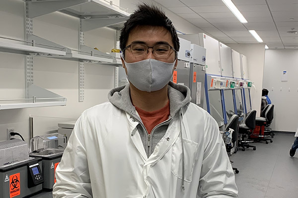 Masahiro standing in a white lab coat in front of metal shelving.
