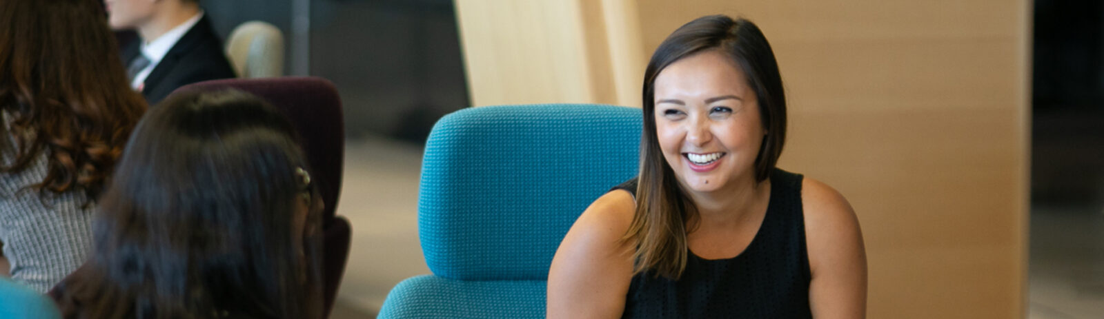 A female student sits in a blue chair and smiles at someone in the foreground.