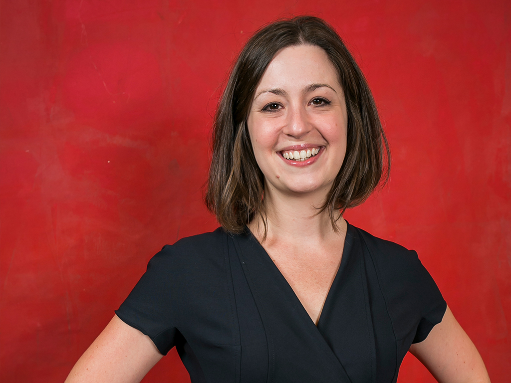 Dina poses in a black top in front of a bright red background.