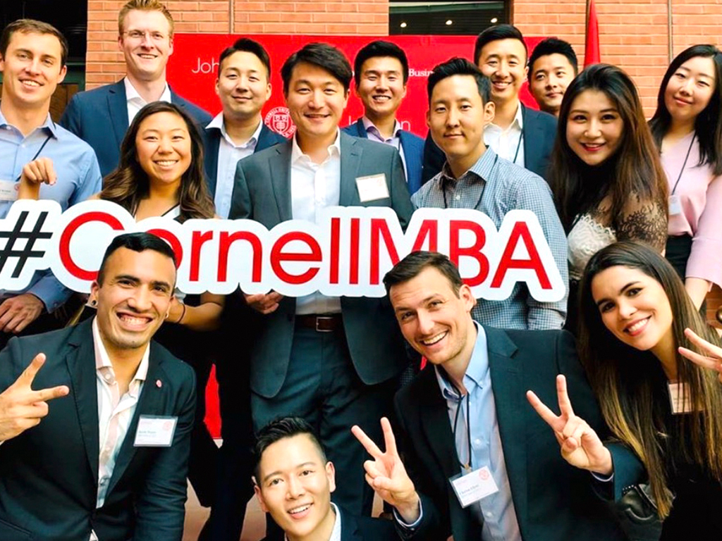 MBA students smile and pose with a sign reading “#CornellMBA.”
