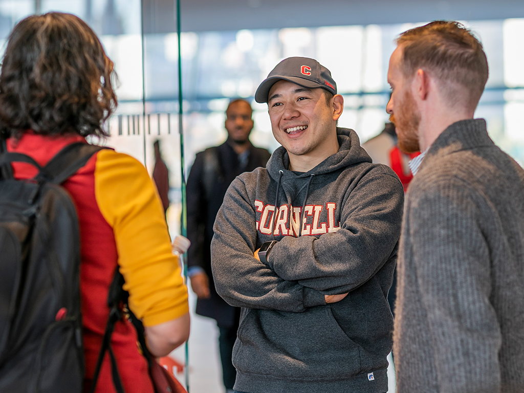 A student in a Cornell hoodie and baseball hat stands smiling with his arms crossed speaking with two other people.