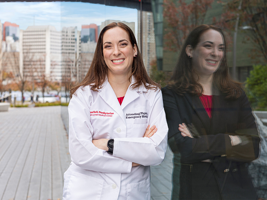 A woman in a lab coat leans against a wall which shows her reflection in business attire.