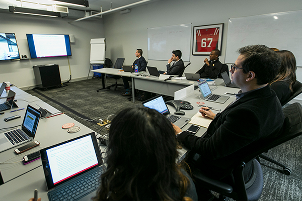 Students sit at a u-shaped table with their laptops watching live instruction on large TVs.