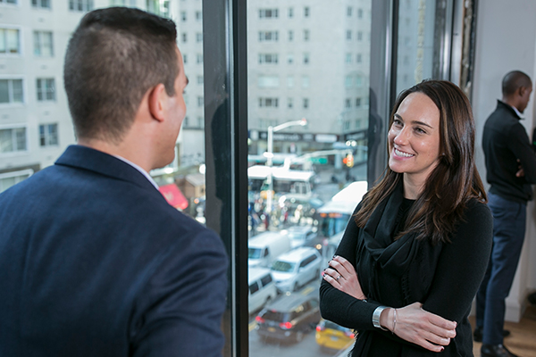 Two students stand and chat with each other in front of a wall of windows with NYC visible outside.