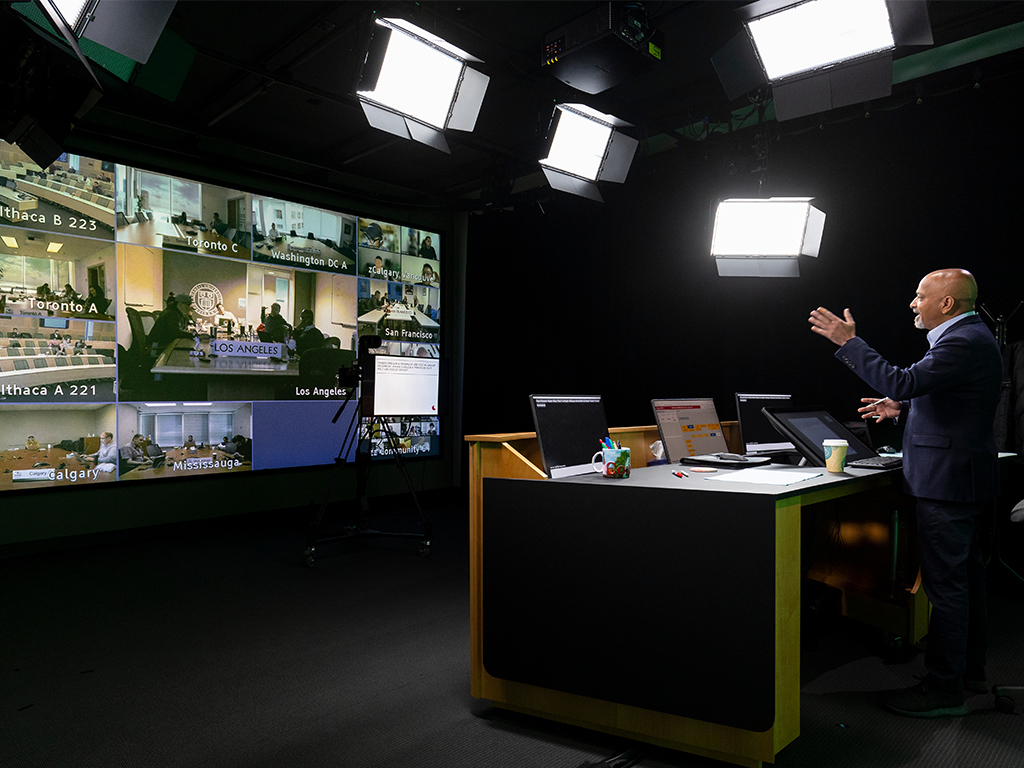 A professor stands at a desk in a TV studio broadcasting him teaching to boardrooms across the Americas.