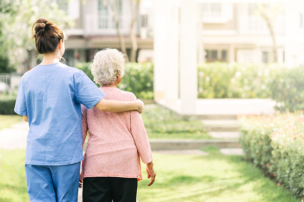 Photo taken from behind of a young woman in scrubs with her arm around an elderly, white-haired woman walking through green gardens with a large home or building in the background.