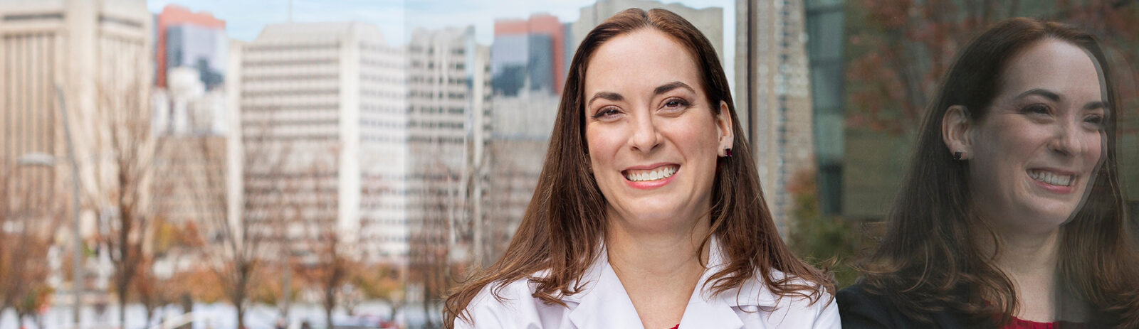 A woman in a lab coat leans against a wall which shows her reflection in business attire.