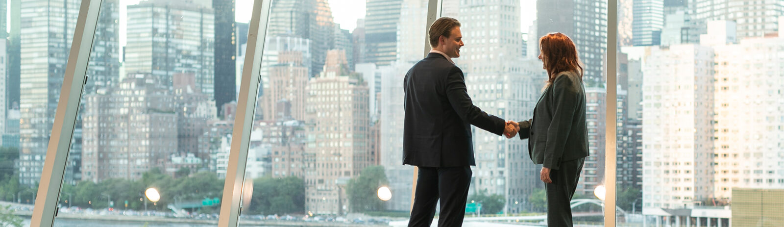 A man in a suit shakes the hand of a woman in a suit in front of a wall of windows with the NYC skyline visible in the background.