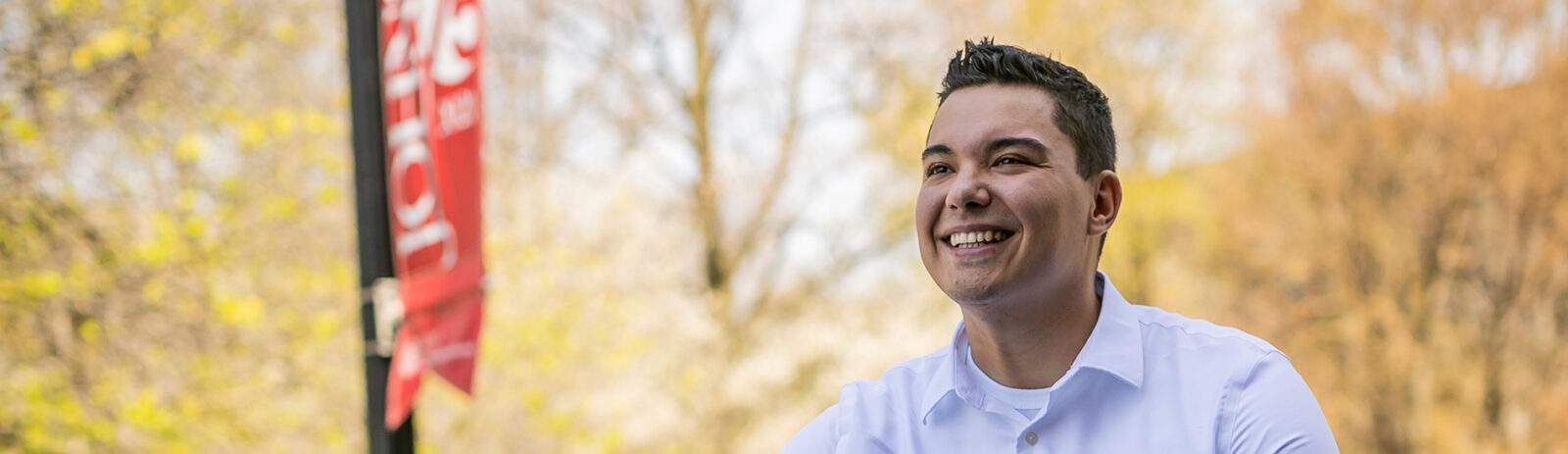 A man in a button up shirt smiles at something out of frame with trees and a Johnson School flag in the background.