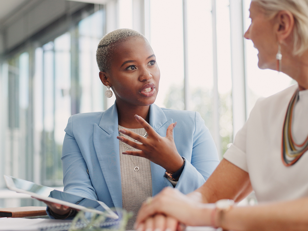 A woman in a blazer speaks holding an iPad to another woman.