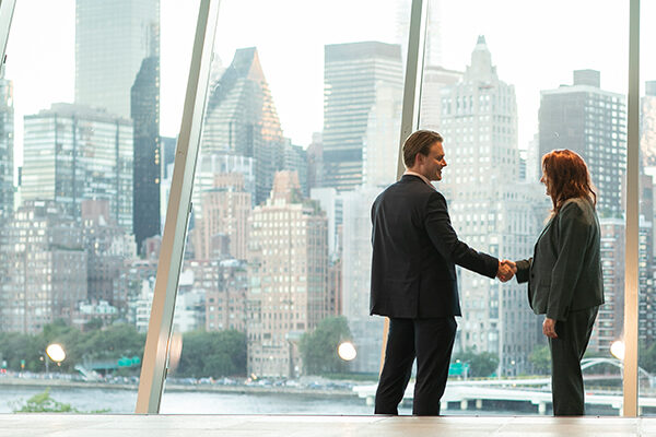 Two people shaking hands in business attire in front of a wall of windows with New York City skyline in the background.