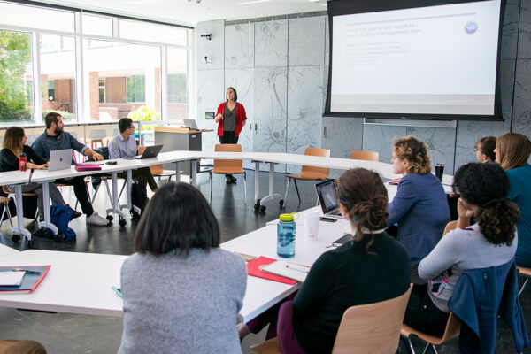 A presenter stands in front of a room next to a projector screen as people look on from a circle of tables.