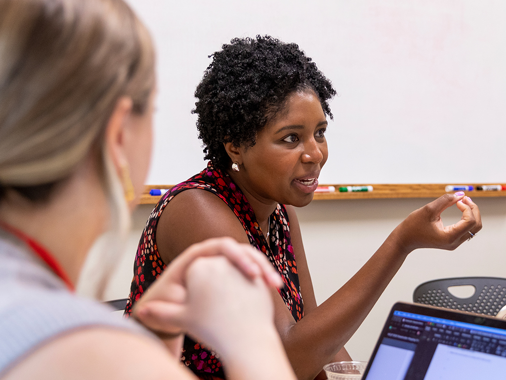 A woman speaks with her hands with a whiteboard in the background.