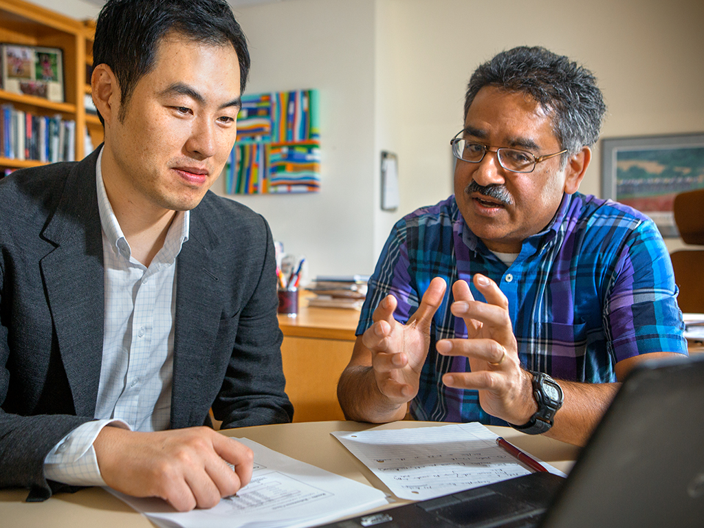 A professor of works with a PhD candidate seated at a table looking at a laptop together.