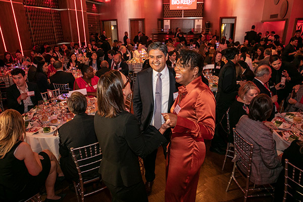 a room filled with people sitting and conversing at dinner tables, with 2 women and one man standing, laughing and talking, in the foreground.