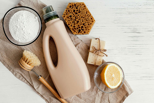A picture of eco- friendly cleaning supplies including a bowl of baking soda, half a lemon, a sponge, soap, scrub brush and a beige bottle of cleaner.