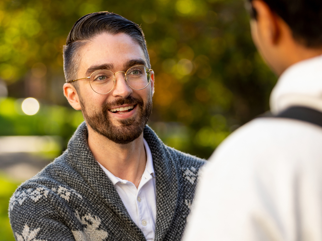 A man in a cardigan and glasses smiles at a person in the foreground.