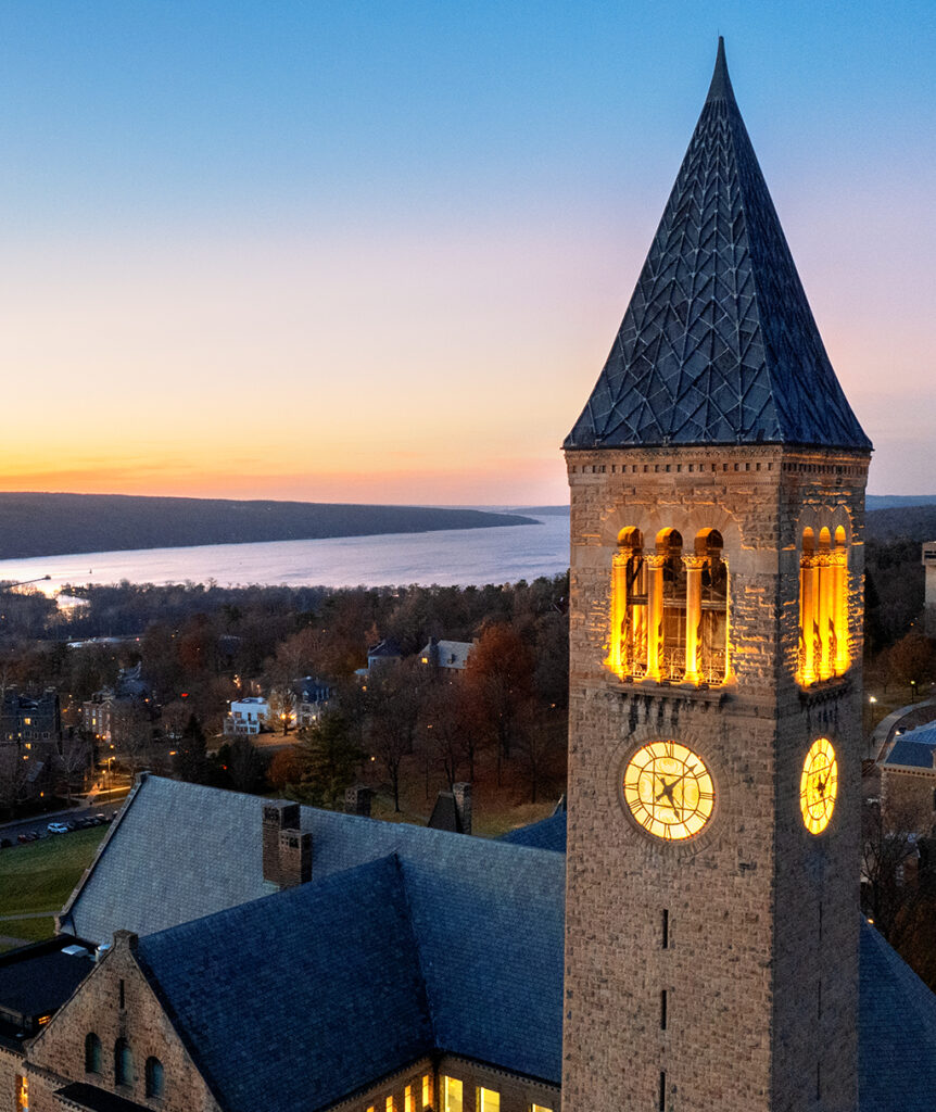McGraw Tower lit up at dusk with Cayuga Lake in the background.