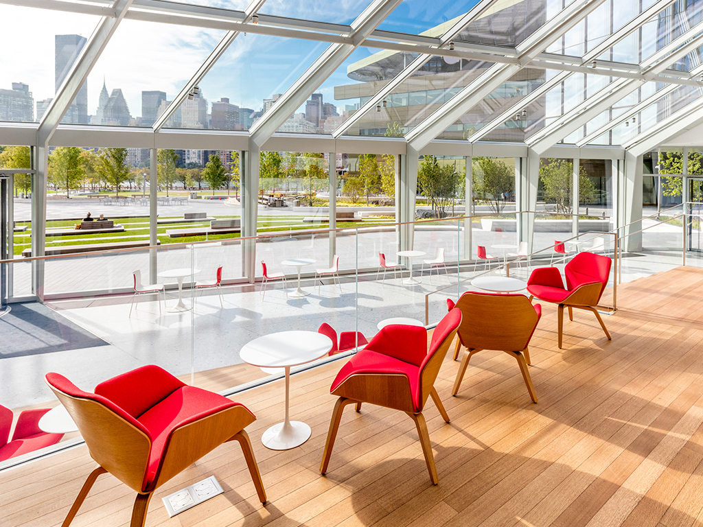 Red soft seating chairs facing a wall and roof of windows with the New York City skyline visible outside.
