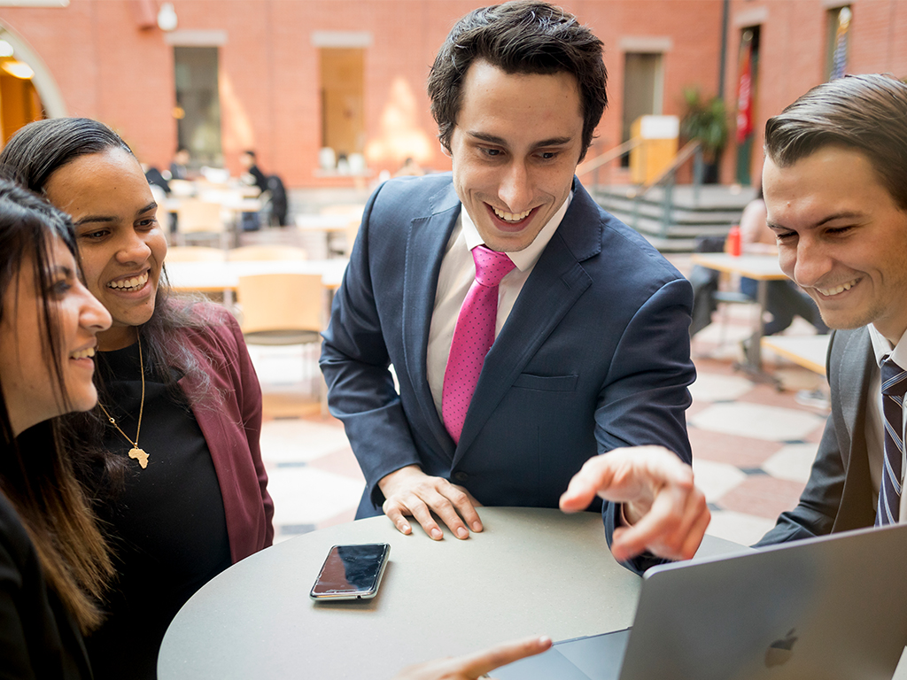 Four people standing around a hightop table smiling and pointing at a laptop.