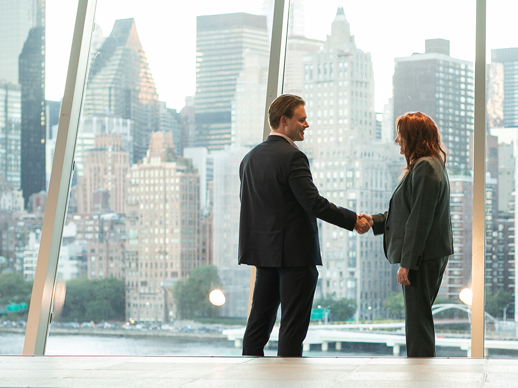 Two people shaking hands in front of a wall of windows with the NYC skyline visible outside.