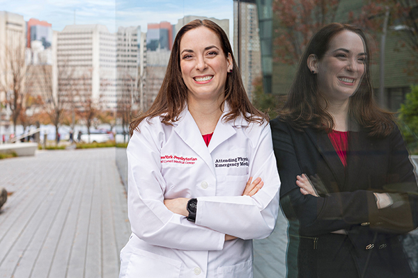 A woman in a lab coat leans against a wall which shows her reflection in business attire.