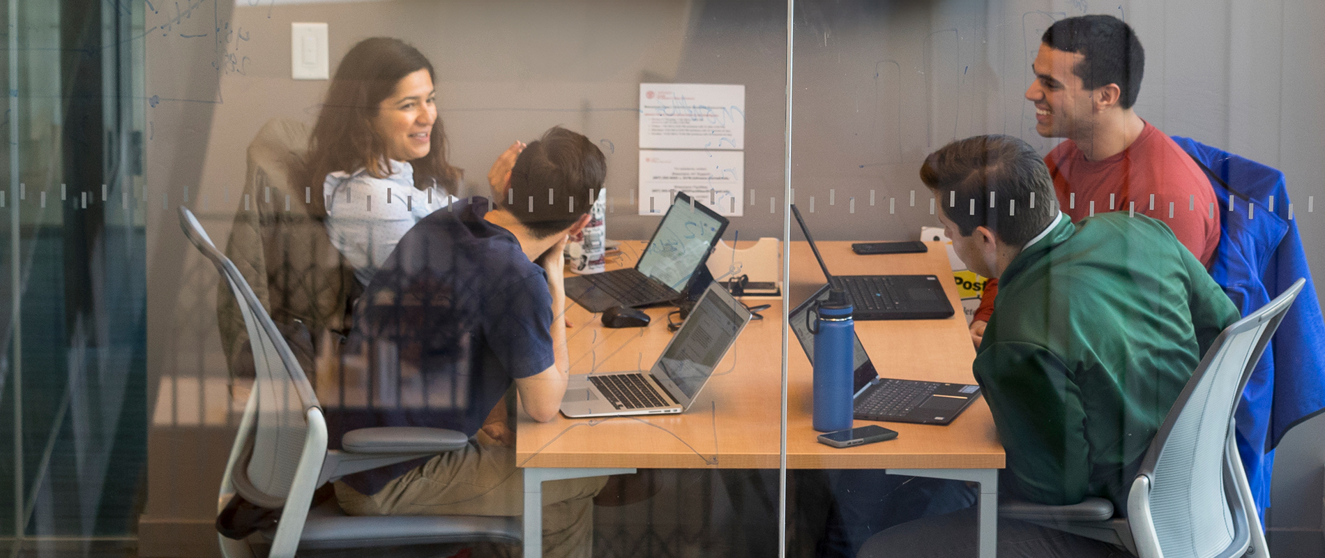 Four people sit at a table with laptops open in front of them.