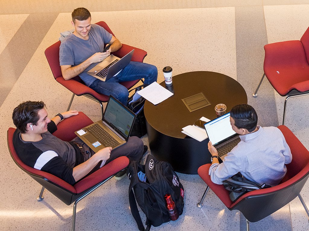 Three students sit in a soft seating area with their laptops smiling at each other.