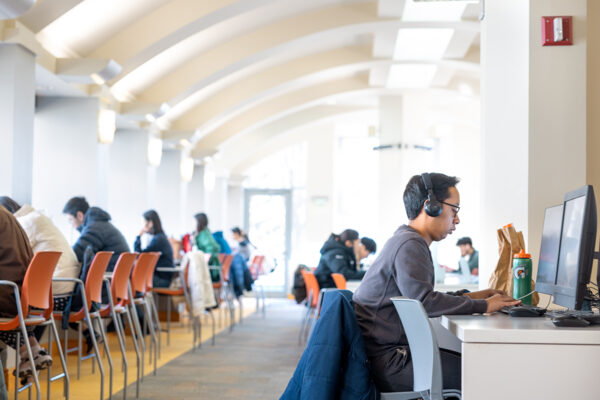 A student sits at a table with a desktop computer wearing headphones with other students working visible in the background.