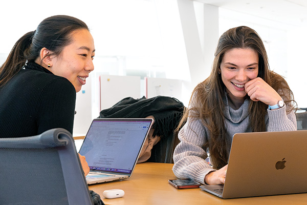 Two students sit at a table with laptops open in front of them.