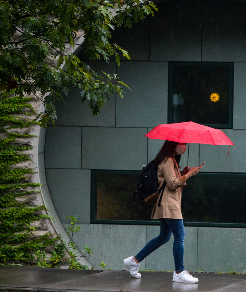 A student walks on the sidewalk in a mask holding a red umbrella.