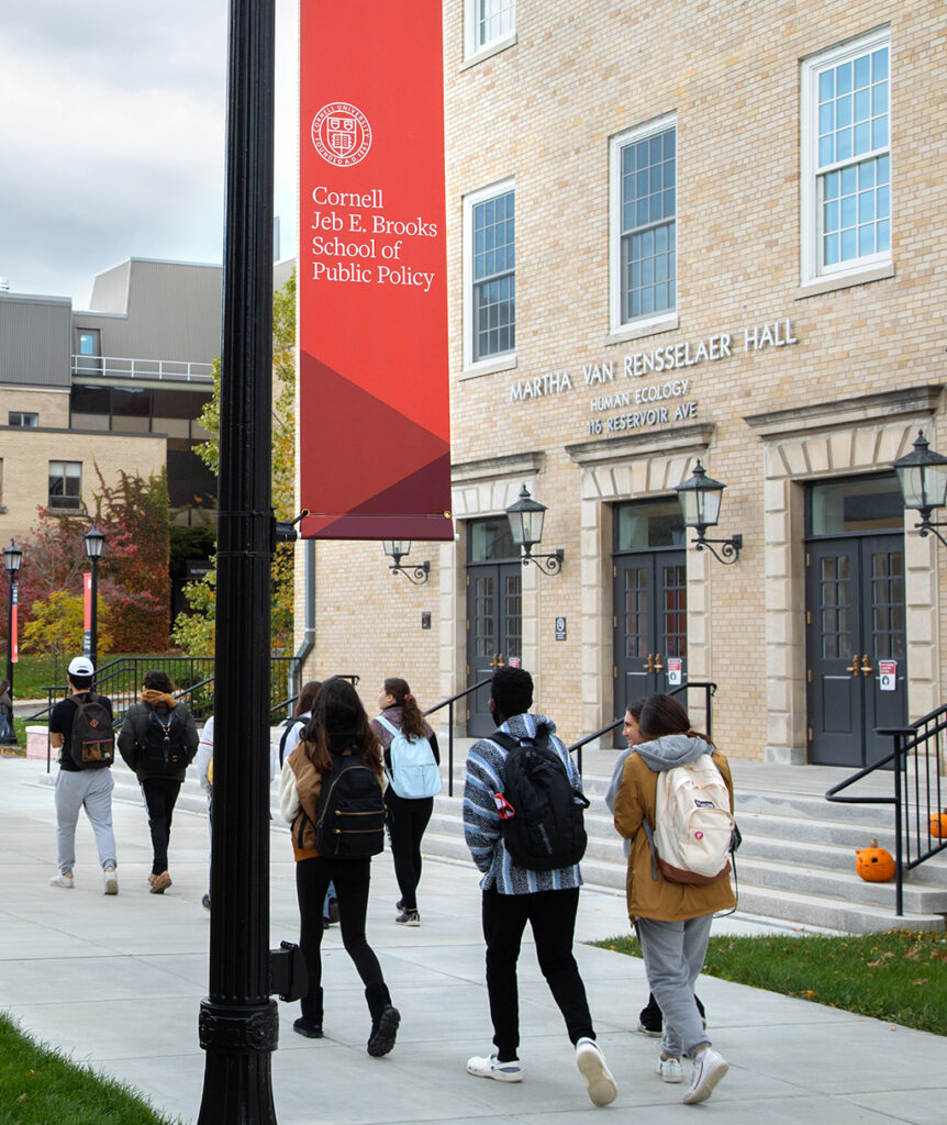 Students walk in front of a Human Ecology building with a Cornell Jeb E. Brooks School of Pubnlic Policy sign in the foreground.