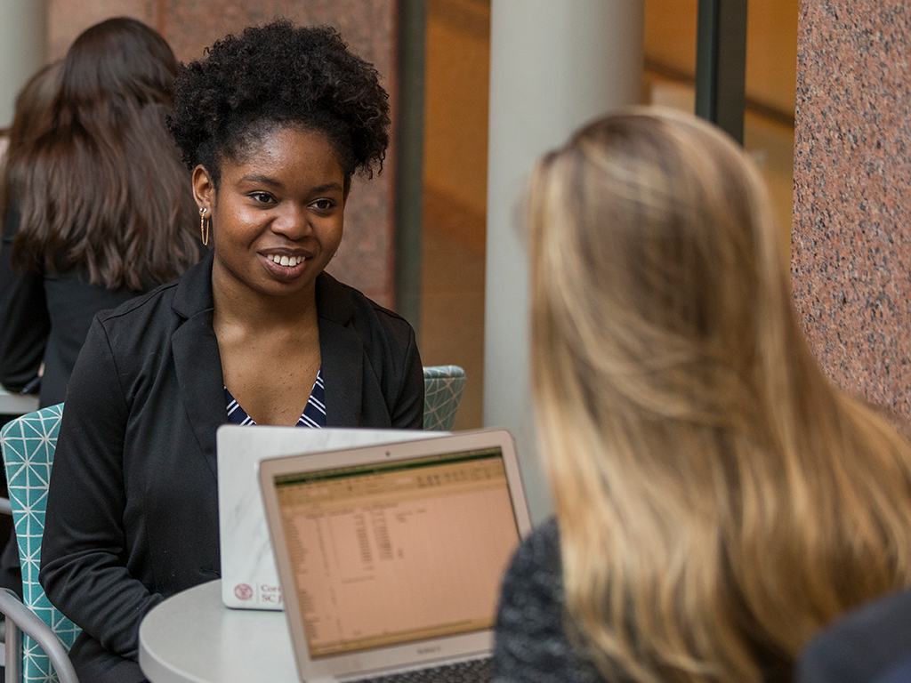 Two women sit at a table with laptops open in front of them.