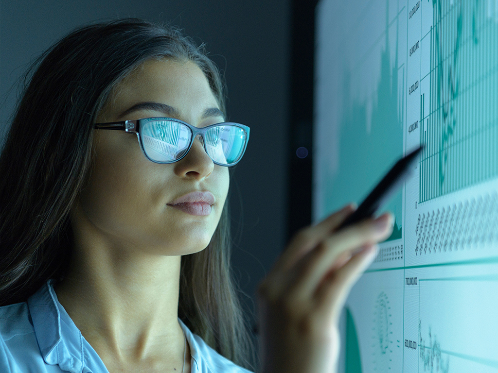 A woman wearing glasses uses a pen to point at charts on a digital screen.