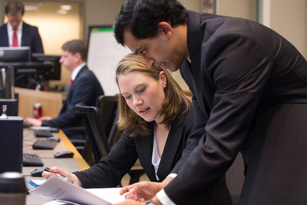 A woman in business attire sits in front of a computer holding paper and discussing them with a man standing next to her in a suit.