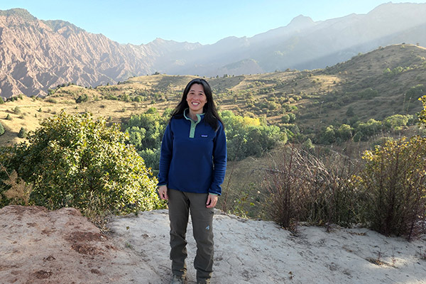 a woman dressed in hiking boots and pants and a fleece standing in a mountainous area surrounded by grean meadows, shrubs, and jagged mountains in the background.