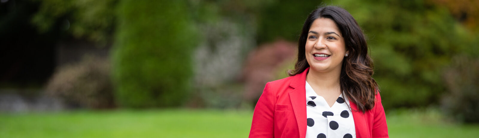 Young woman in red business jacket and black-and-white polka dot shirt smiles off-camera in a green field with a row of trees behind, out of focus