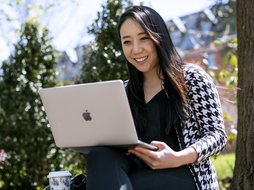 A woman sits outside on an Apple laptop with trees visible in the distance.