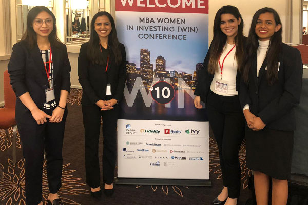 Four women in business attire pose next to a “MBA Women in Investing Conference” sign.